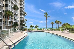 A swimming pool with a clear blue sky, palm trees, and a high-rise building in the background.