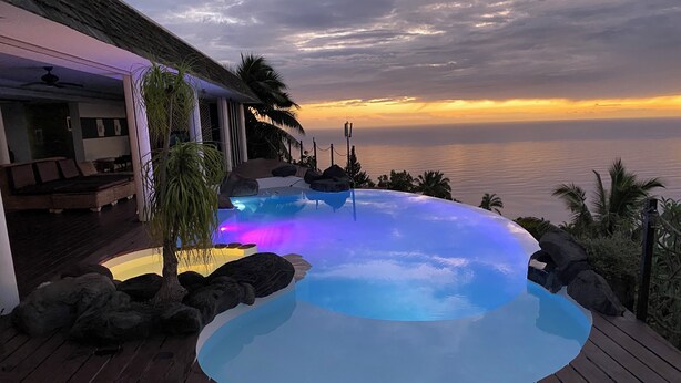 Une piscine à bordures courbes, entourée de rochers et de palmiers, offrant une vue sur l’océan au coucher du soleil.