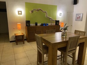 Een eethoek met een houten tafel en stoelen, een lamp, een speaker en een groene wand met een takdecoratie.