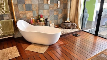 A modern bathroom with a freestanding bathtub, wooden flooring, stone wall tiles, and a view through a glass door.