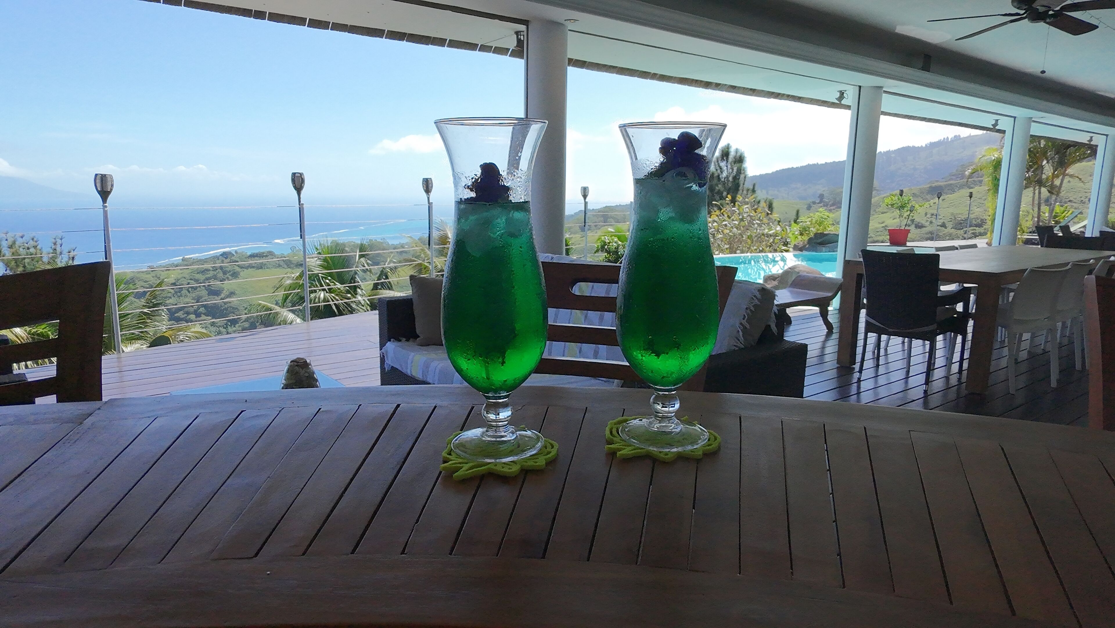 Deux grands verres remplis d’une boisson verte sont posés sur une table en bois, offrant une vue sur un paysage montagneux.