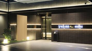 A modern hotel entrance with a glass door, illuminated signage, and a small outdoor seating area with plants.