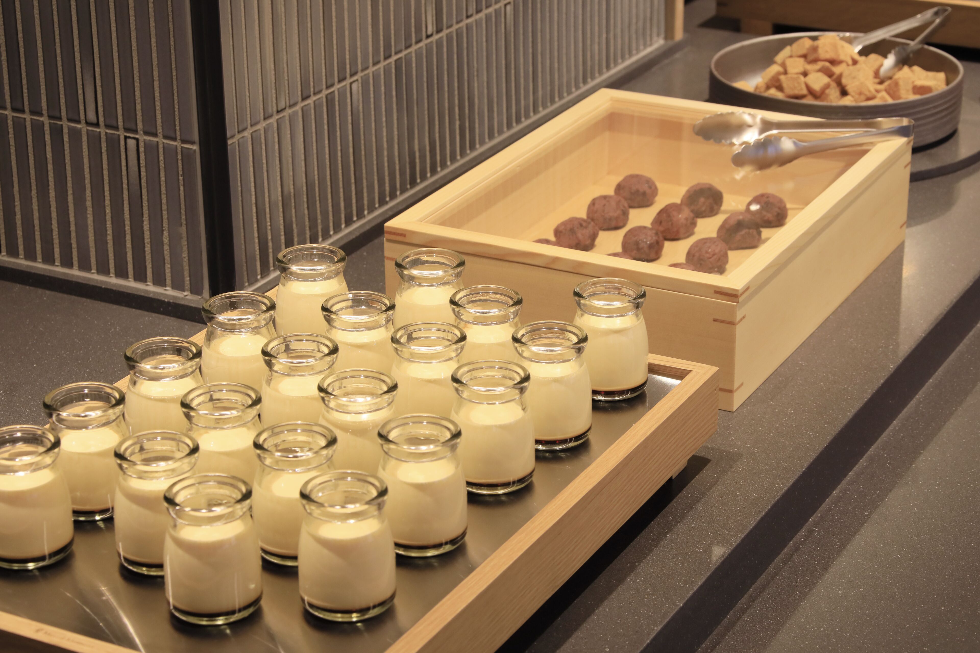 A tray of small glass jars with a creamy substance, a wooden box with round objects, and a bowl with cubes and a spoon.