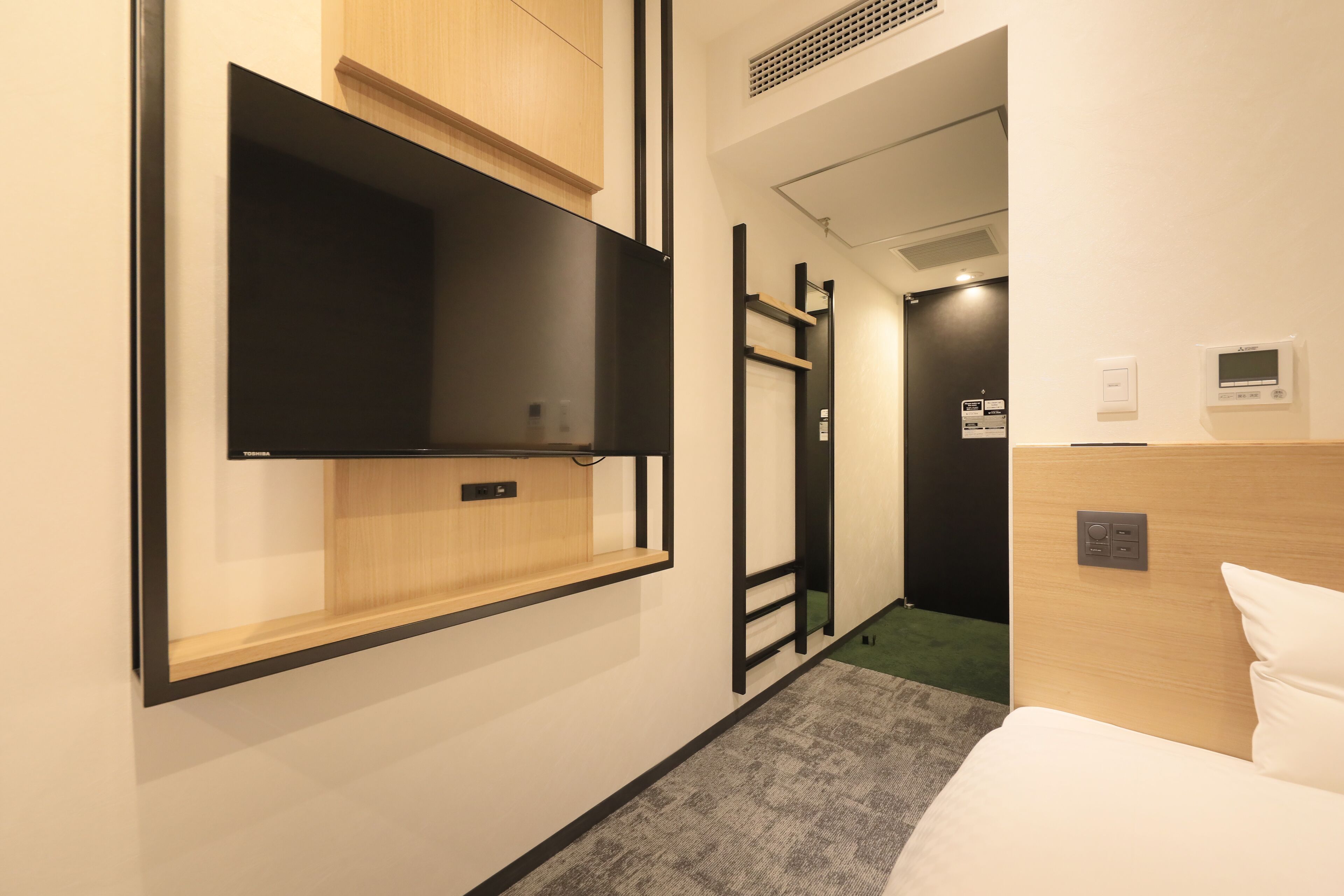 A hotel room with a bed, a TV mounted on the wall, a wooden cabinet, a control panel, and a door leading to another room.