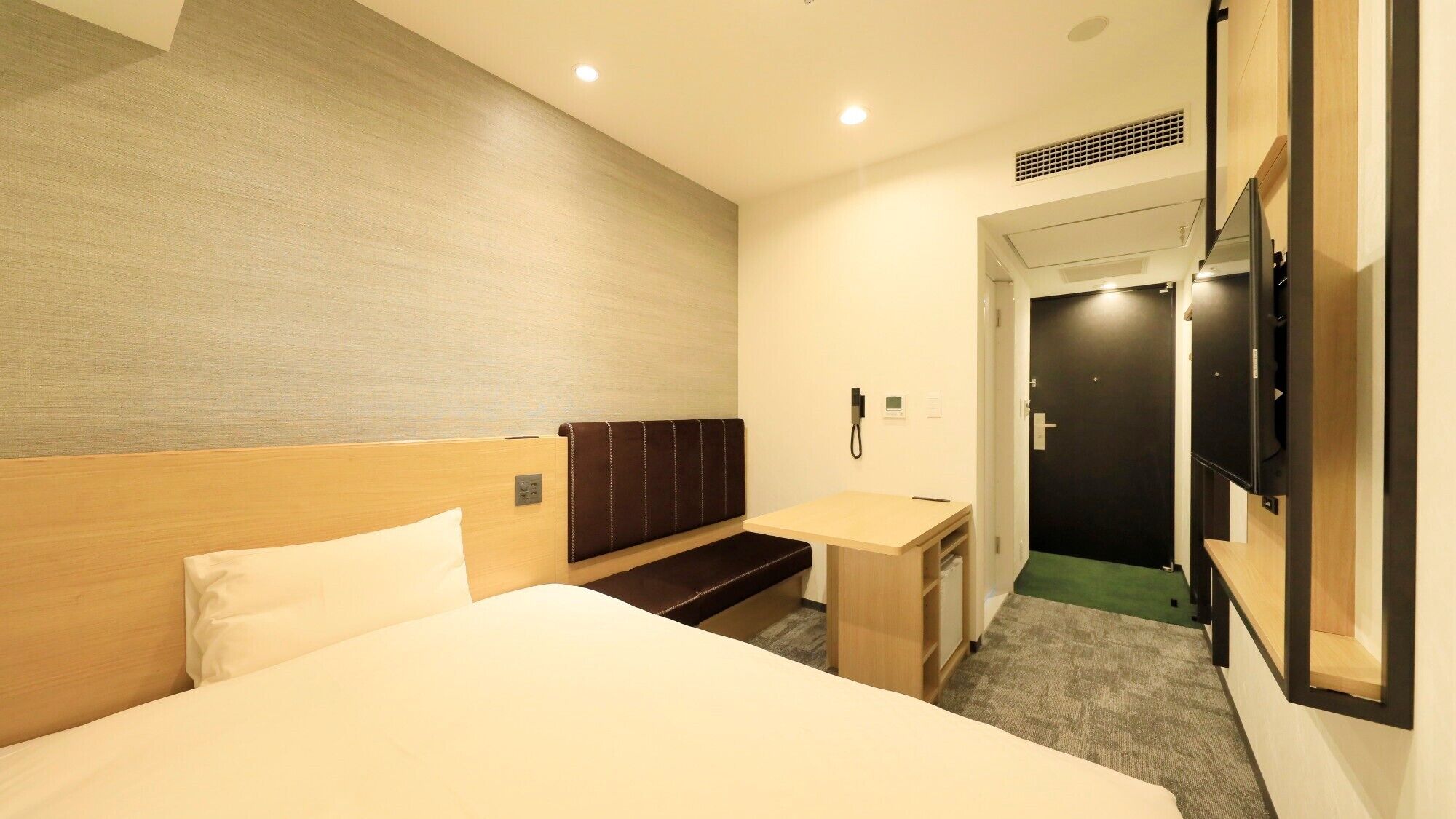 A hotel room with a bed, a desk, a bench, a TV, and a door.