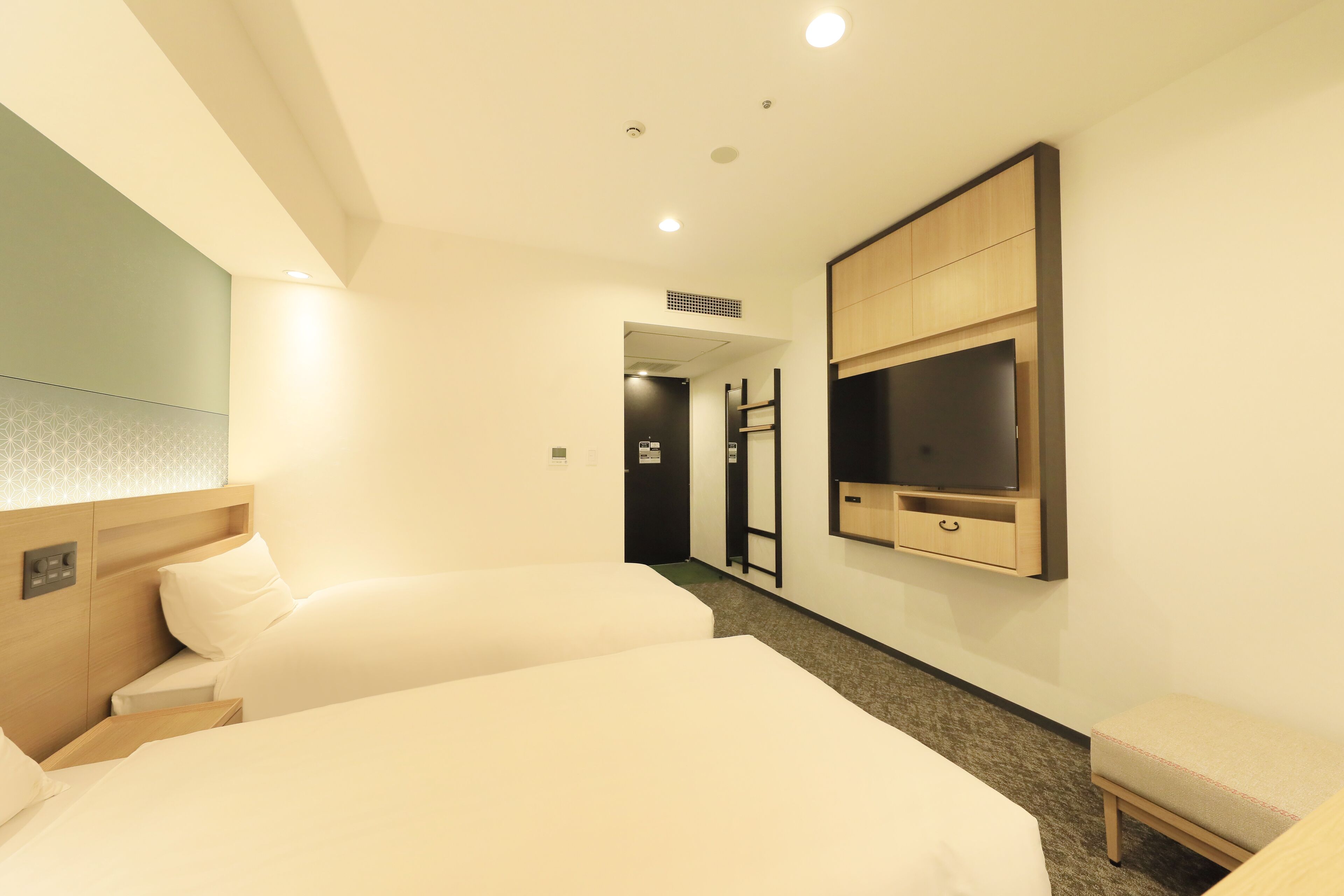 Habitación de hotel con dos camas, un televisor montado en la pared, un pequeño banco y una puerta que da a un baño.