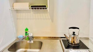 A compact kitchen with a stainless steel sink, a kettle, and a small shelf above the sink holding dishes and a hairdryer. - TIME SHARING STAY ikebukuro (Tokyo)