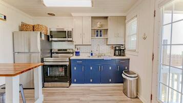 Cocina compacta con armarios blancos y azules, electrodomésticos de acero inoxidable y una ventana que proporciona luz natural.