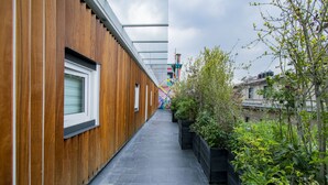 Een smalle, buitenste gang met houten lambrisering aan de ene kant en een vloer van zwarte tegels. Aan de rechterkant staan plantenbakken met groen.