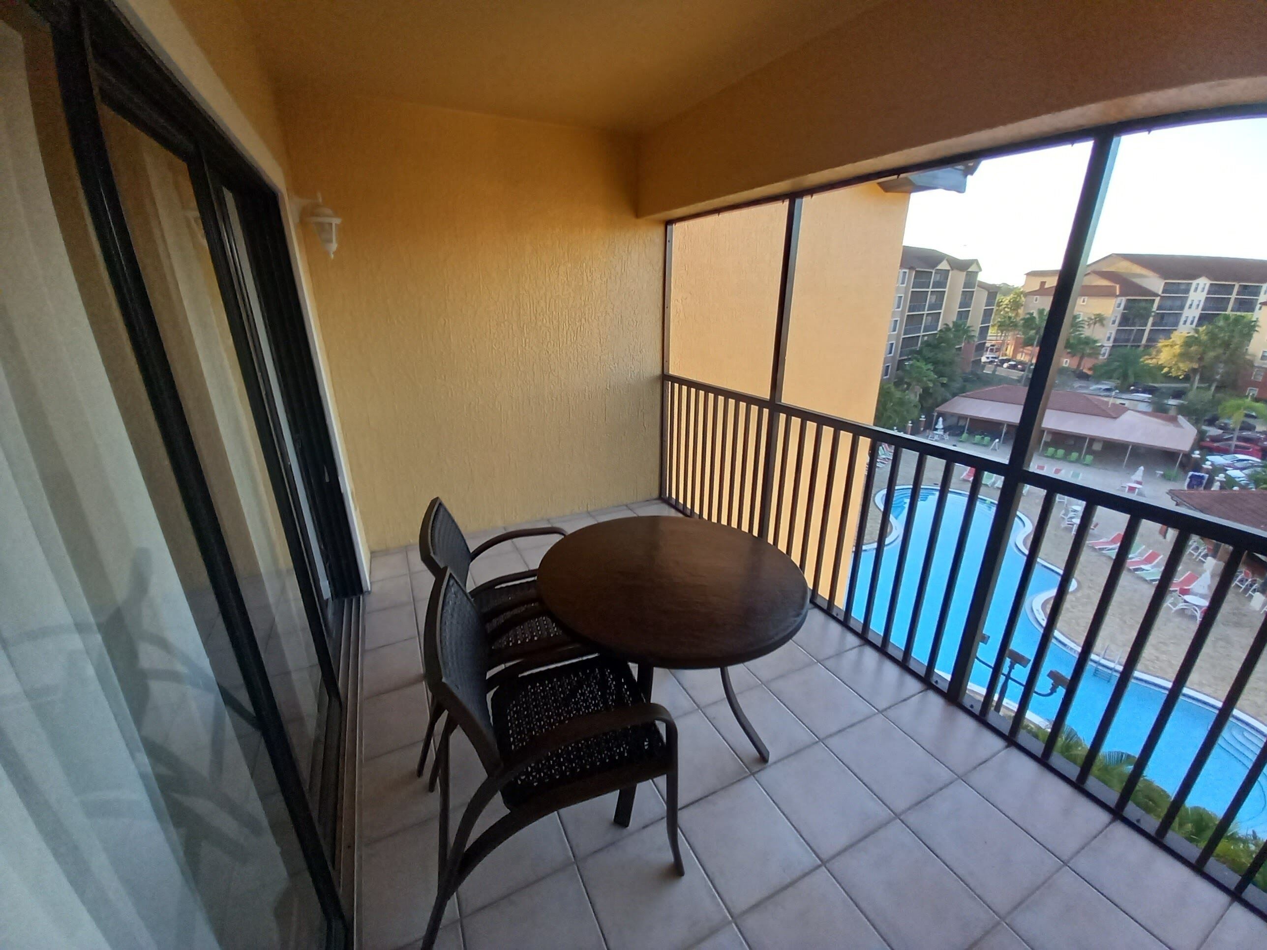 A balcony with a table and chairs, a view of a swimming pool, and buildings in the background.