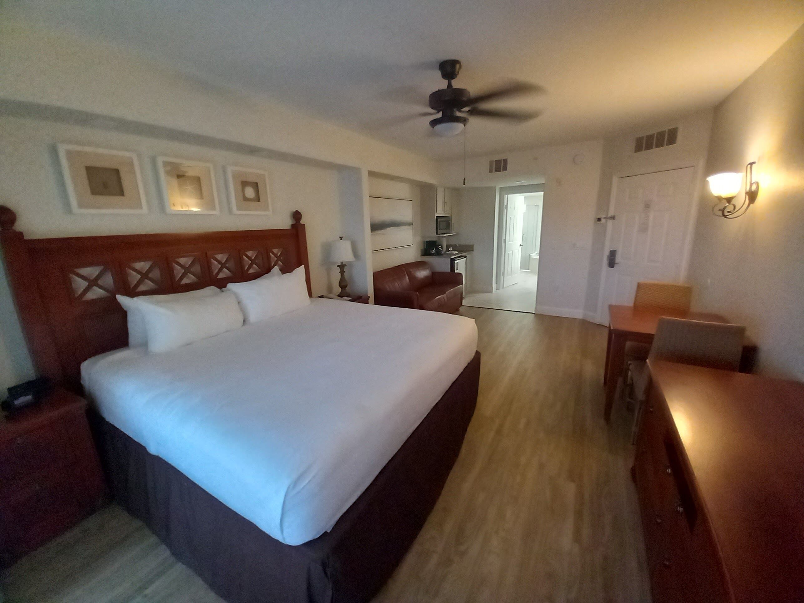 A hotel room with a large bed, a desk, a chair, a ceiling fan, and a door leading to another room.