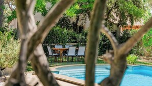 Una piscina con cubierta de madera y sillas, rodeada de abundante vegetación y un edificio al fondo.