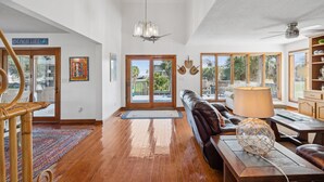 A spacious living room with a wooden floor, a leather sofa, a chandelier, and a view through large windows. - 1511 Forrest Ave - Sea Caught The Katy - Private Pool- Ocean Views-Best Surf at Washout (Folly Beach)