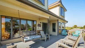 A covered outdoor deck with wooden furniture, including a swing chair and lounge chairs, and large windows offering views of the surrounding area. - 1511 Forrest Ave - Sea Caught The Katy - Private Pool- Ocean Views-Best Surf at Washout (Folly Beach)