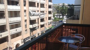 A balcony with a metal table and chairs, overlooking a multi-story building with balconies and a street below.