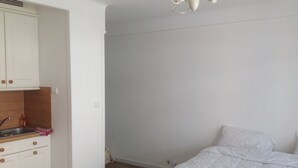 A bedroom with a bed, a white wardrobe, a kitchen area with a sink, and a ceiling light fixture.