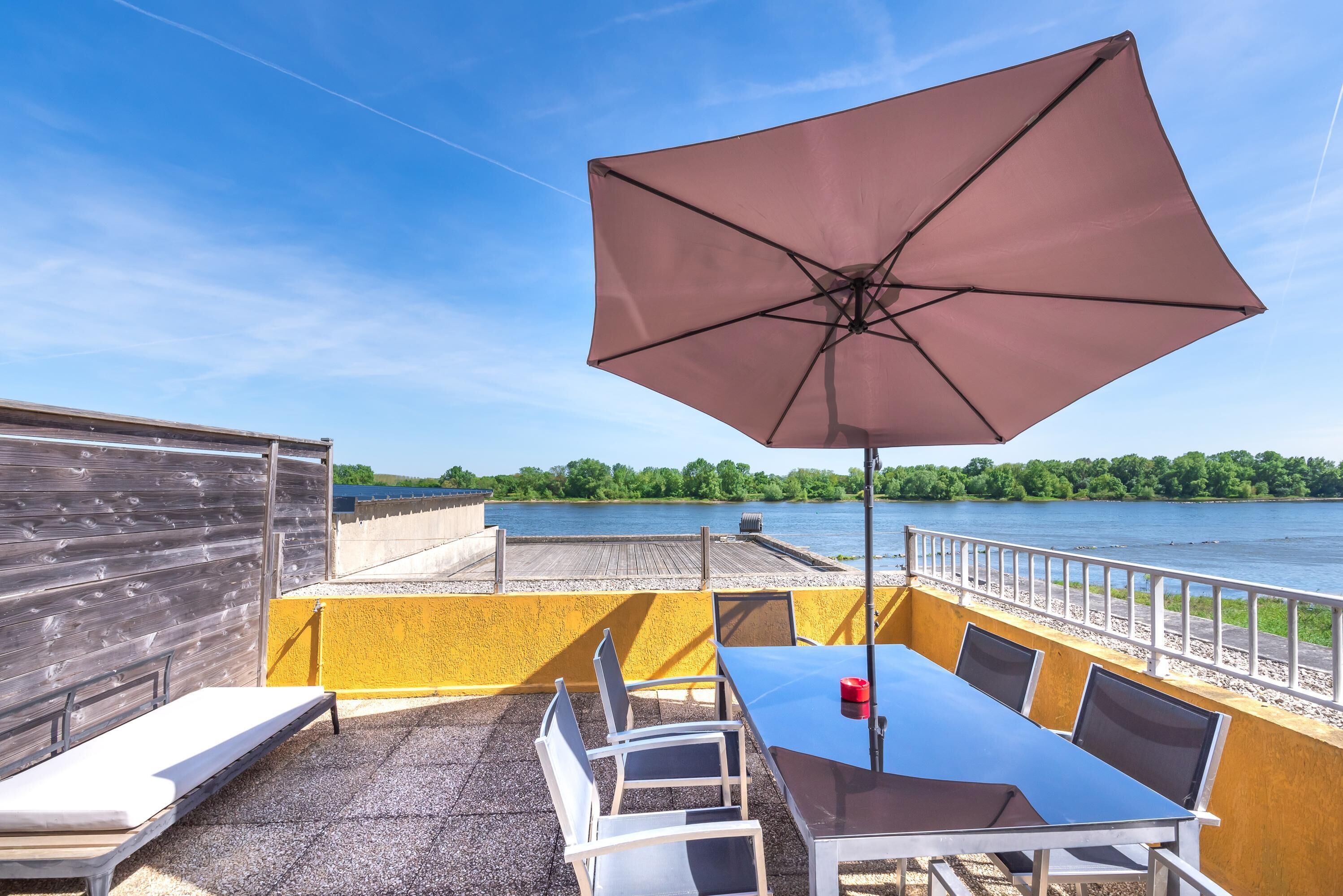 An outdoor patio with a table and chairs, a large umbrella, and a view of a river.