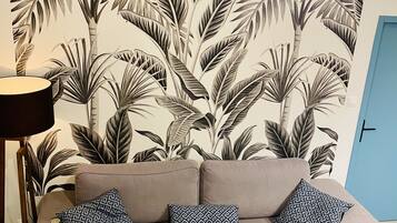 Ein Wohnzimmer mit einer Couch, gemusterten Kissen, einer Pendelleuchte und einer Wandmalerei mit tropischen Blättern.