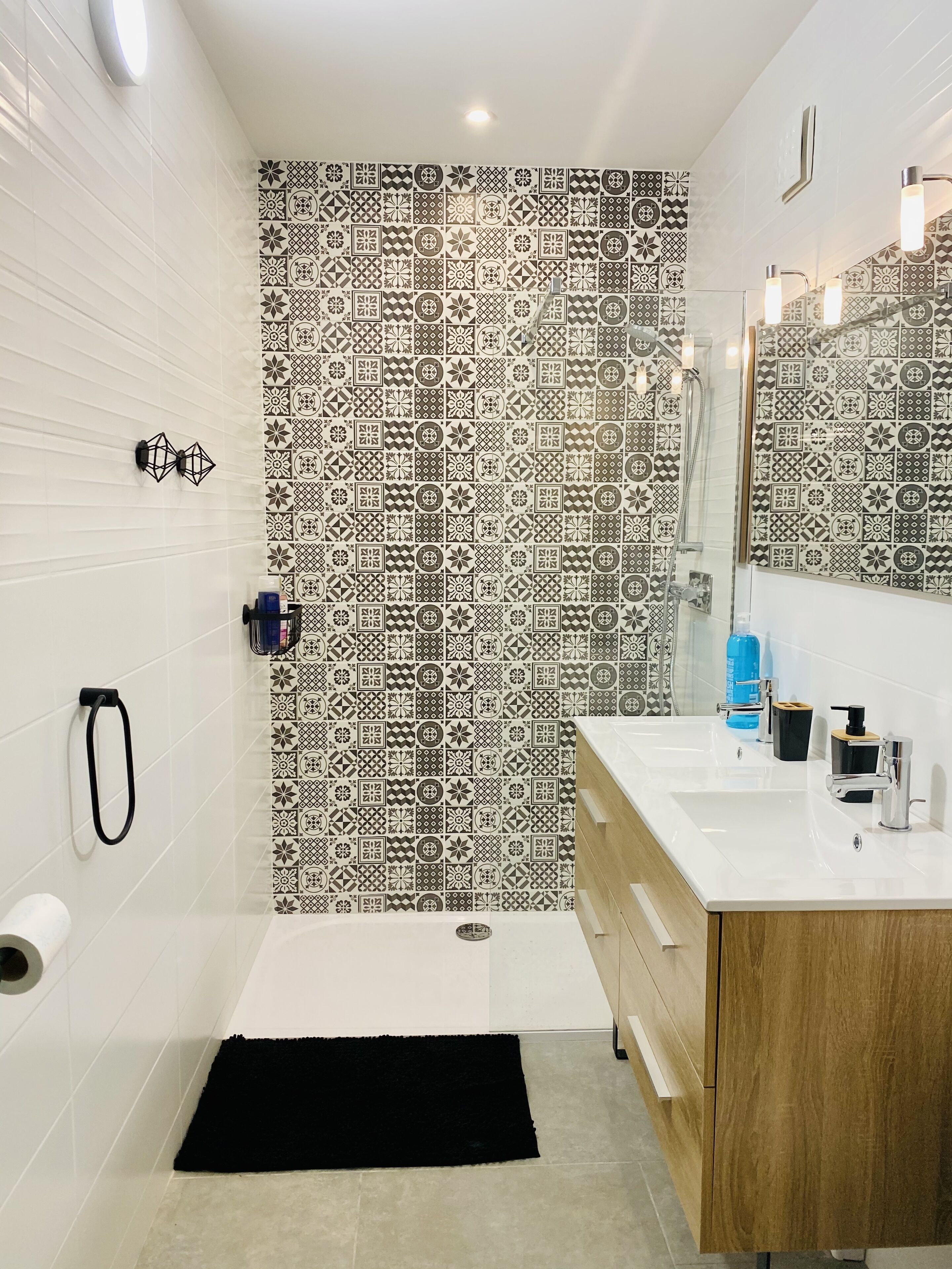 Une salle de bain avec un mur recouvert de carreaux à motifs, un lavabo blanc doté d’accessoires modernes et un tapis noir sur le sol.