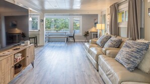 Amplia sala de estar con un sofá de cuero color beige, mueble de entretenimiento de madera y un televisor de pantalla plana grande. Cuenta con una zona de comedor con un banco y una mesa, y vistas a zonas verdes a través de las ventanas.