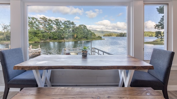 Un comedor con mesa de madera y dos sillas, con vista a un lago que tiene un muelle y vegetación.
