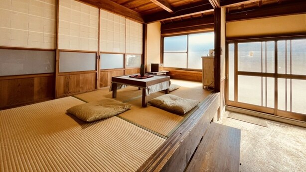 A traditional Japanese room with tatami flooring, a low wooden bed, a table with a chair, and a view through large windows.