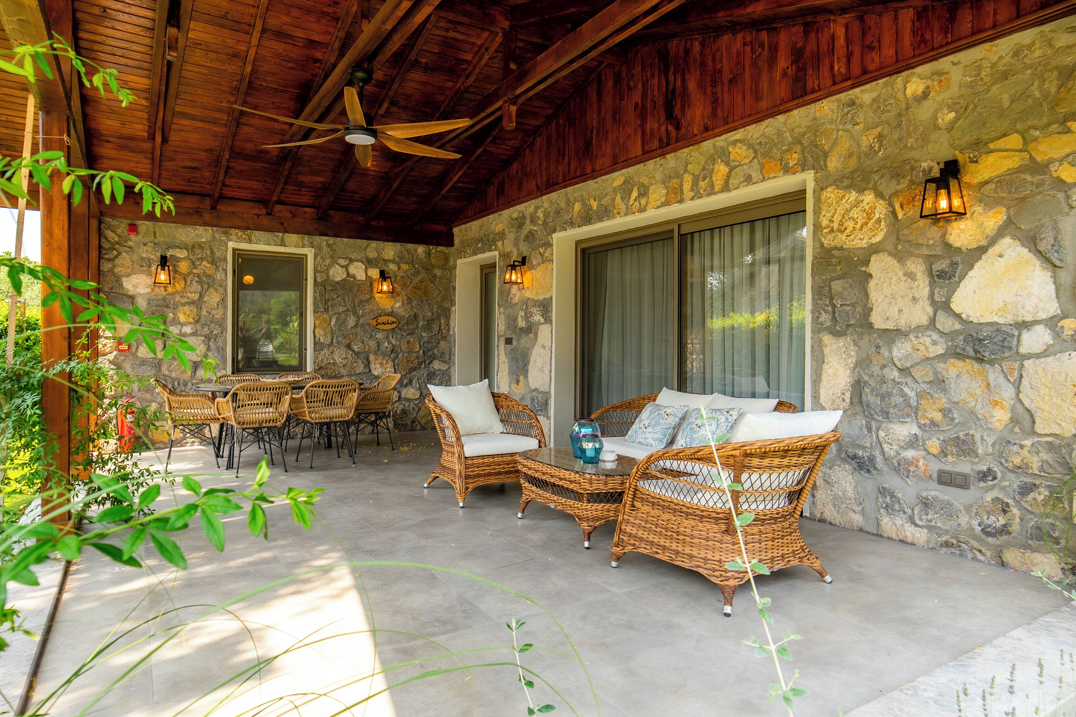 A covered patio area with wicker furniture, stone walls, and a ceiling fan.