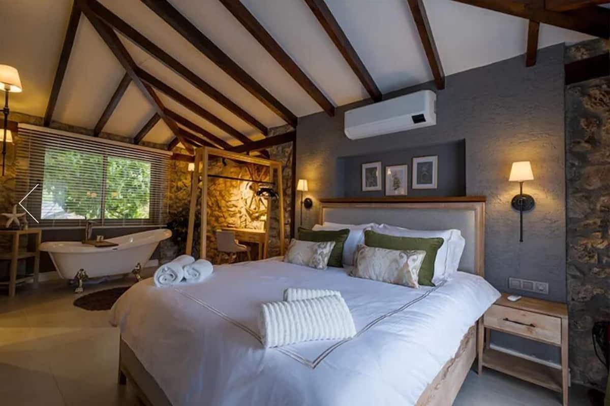 A bedroom with a large bed, a bathtub, a wooden chair, and a stone wall.