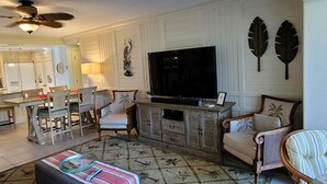 A living room with a television, a dining area with chairs, and a kitchen in the background. - CP 614 Golf Course View Condo - Paradise Vacation Rental in Ocean Village (Fort Pierce)
