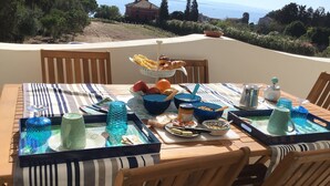 Une table à manger installée à l’extérieur, avec différents plats, une carafe en verre bleue, et en arrière-plan, une vue sur la mer et les collines.