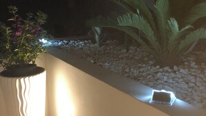 Un espace extérieur moderne agrémenté d’un bac à fleurs blanc, d’une plante en pot et d’un luminaire fixé au mur.