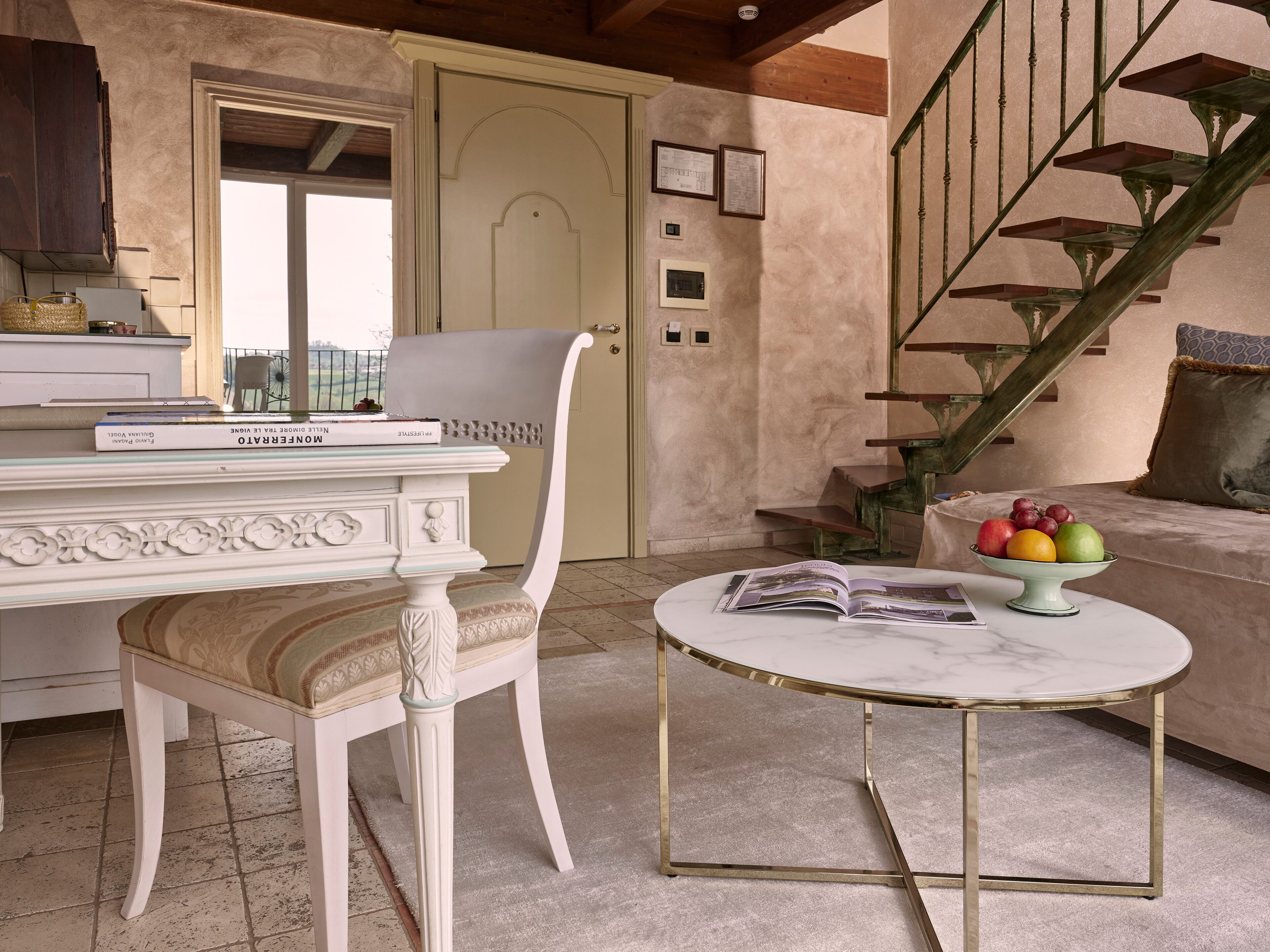 Una habitación con una silla blanca, una mesa redonda con tapa de mármol, una escalera y un frutero en el sofá.