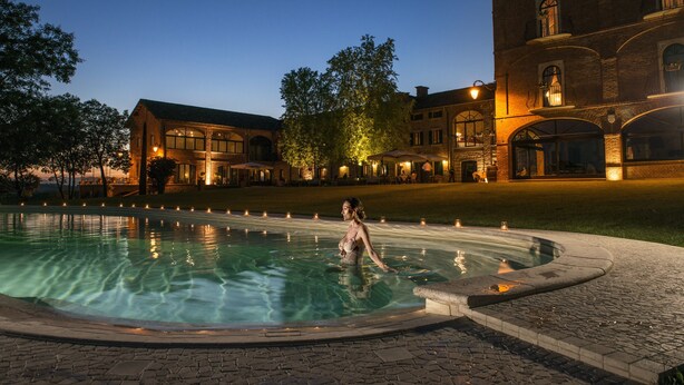 A pool area with a person standing in the water, surrounded by a brick building with multiple windows and a well-lit entrance. - Tenuta Montemagno (Montemagno)