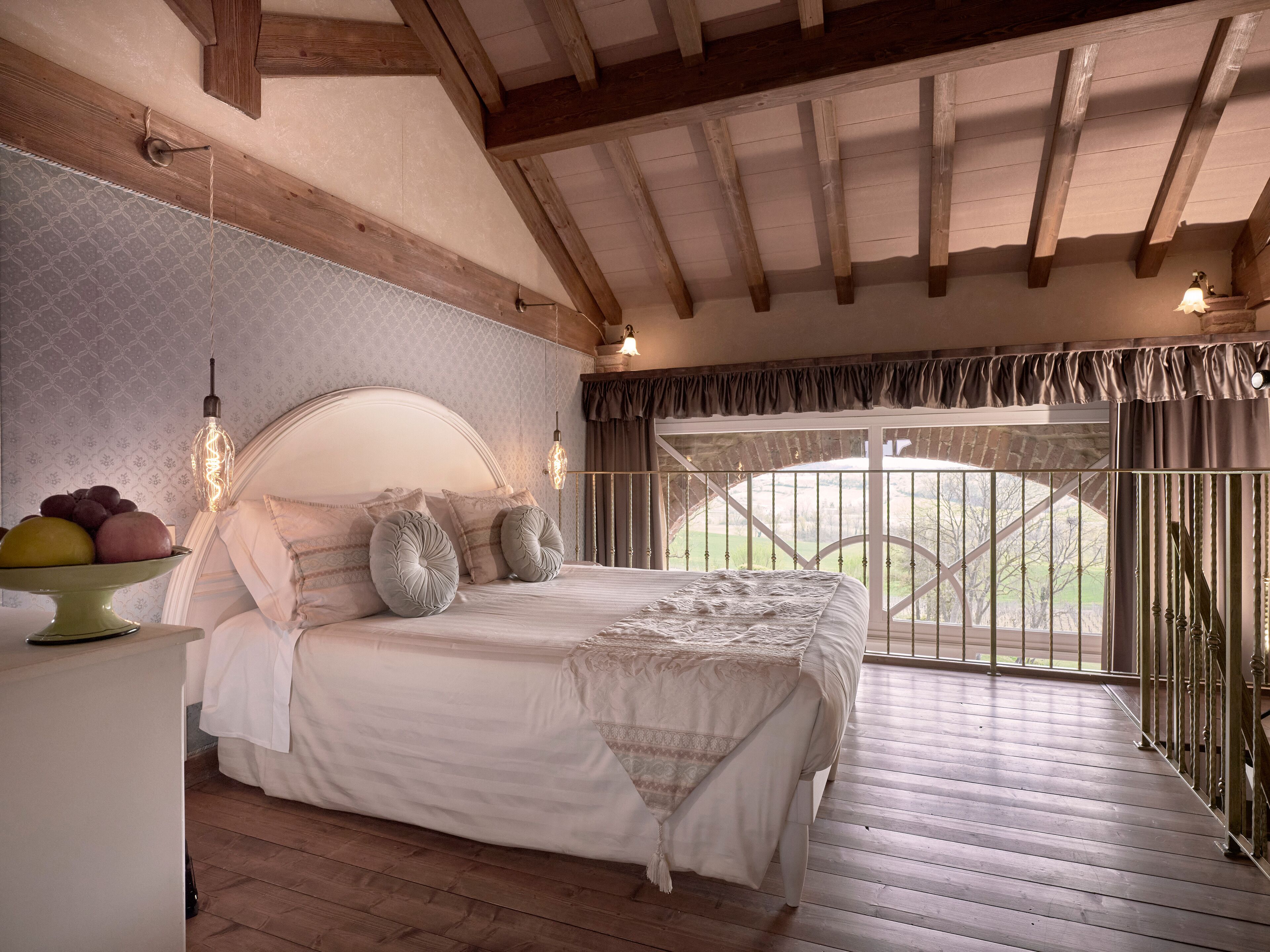 Une chambre avec un plafond en bois, un grand lit doté d’une tête de lit blanche, une table de chevet avec des fruits, et une vue sur l’extérieur par une fenêtre ornée d’une rambarde en métal.