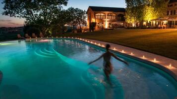 Una piscina al anochecer con una persona nadando, rodeada de árboles y un edificio con terraza.