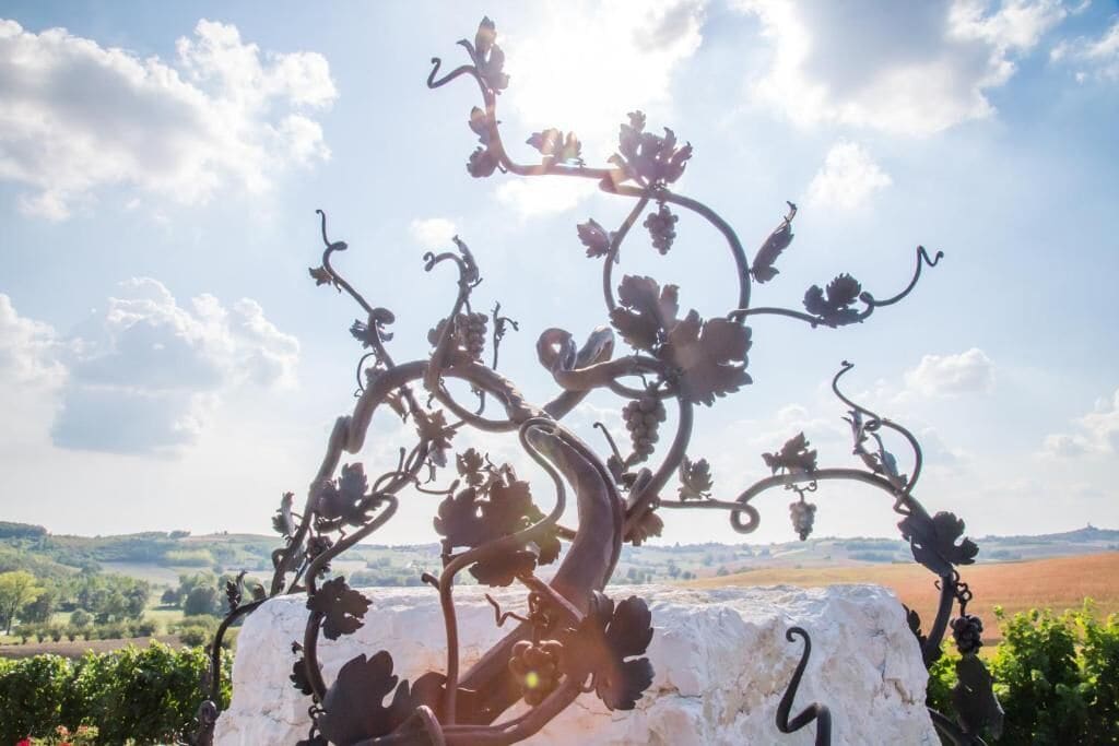 A metal sculpture of a vine with grapes, set against a backdrop of a vineyard and a clear sky.
