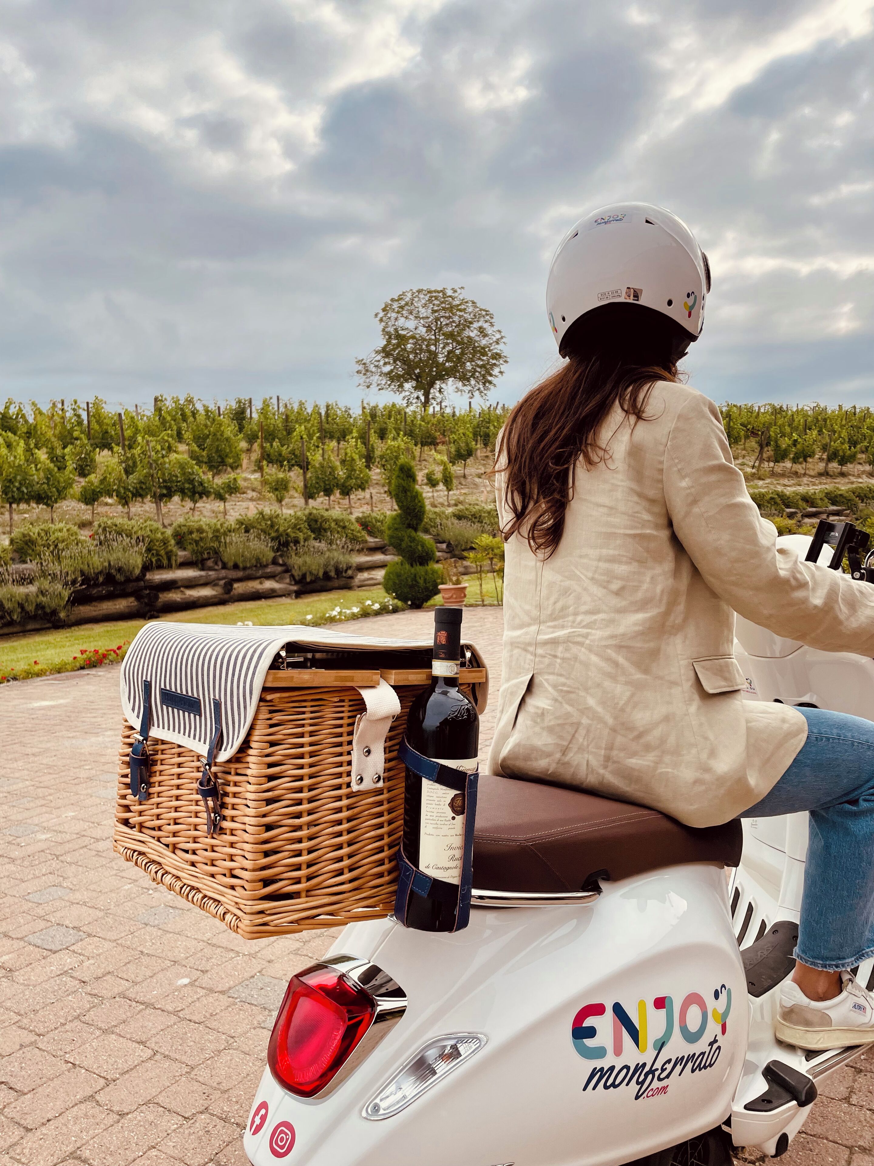 Iemand rijdt op een witte scooter met een rieten mandje en een fles wijn.