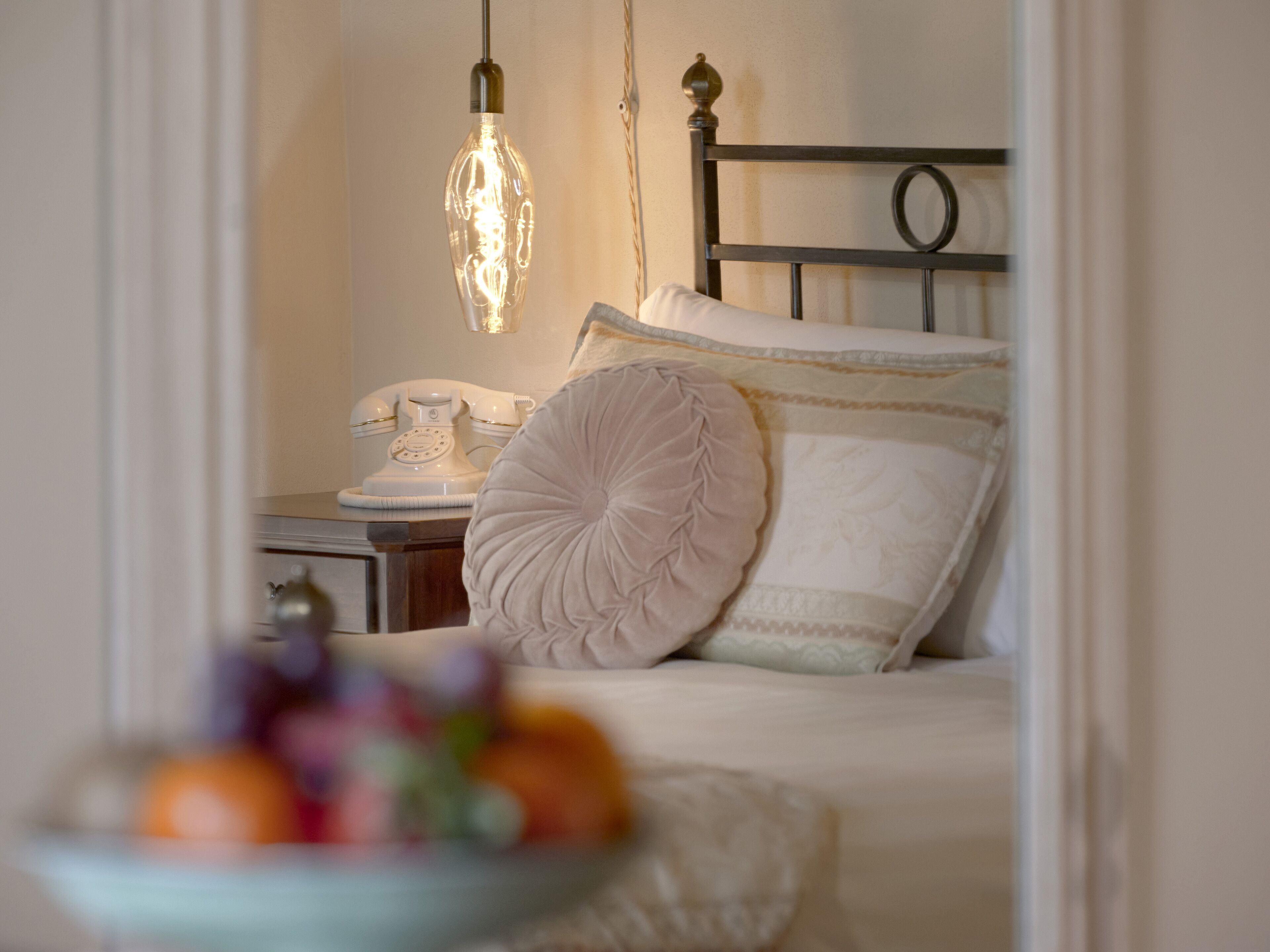 A neatly made bed with white linens and a decorative pillow, a vintage telephone on a bedside table, and a hanging light fixture.