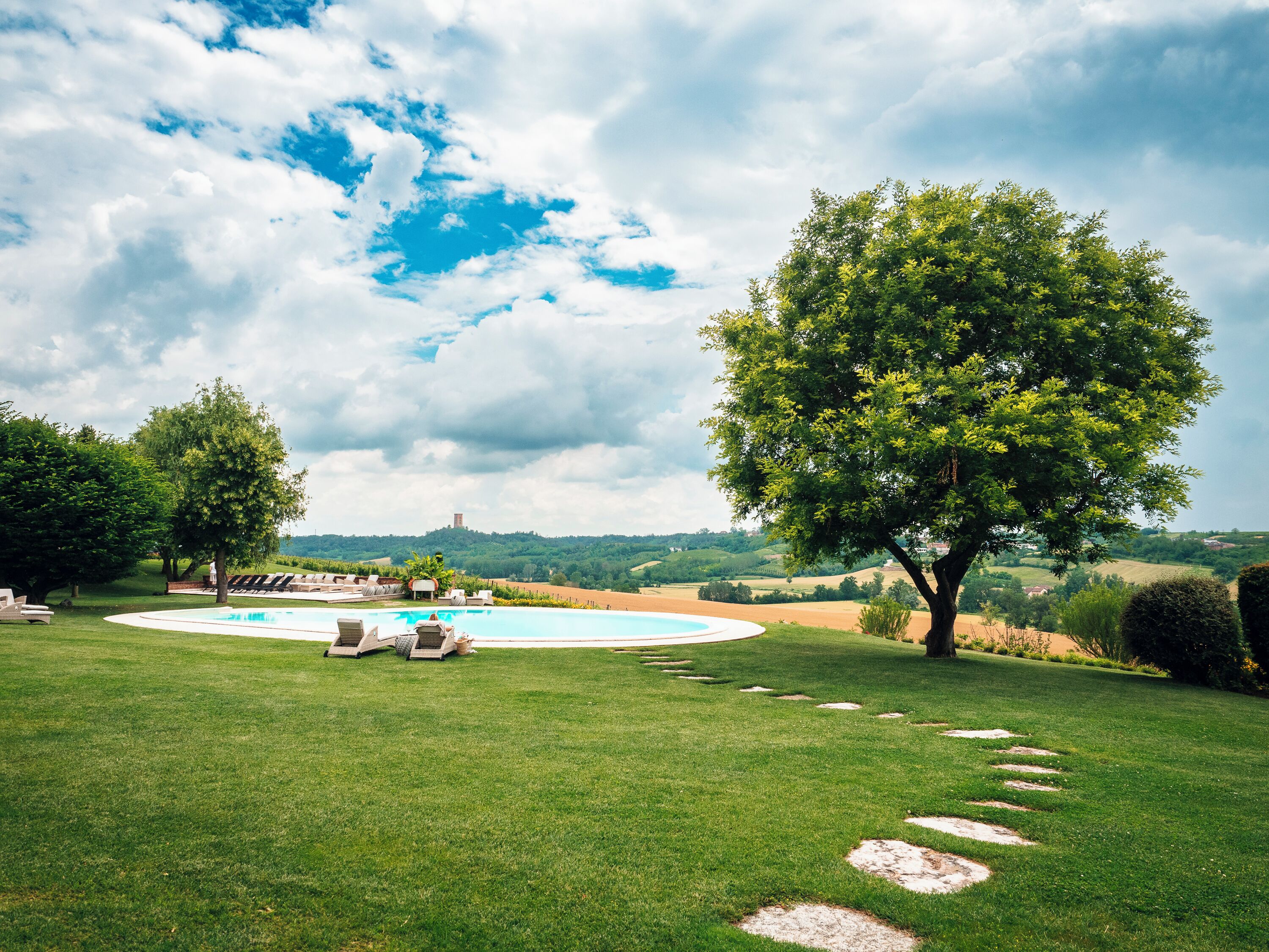 A circular pool with lounge chairs, surrounded by a well-manicured lawn and trees, with a view of rolling hills in the background.