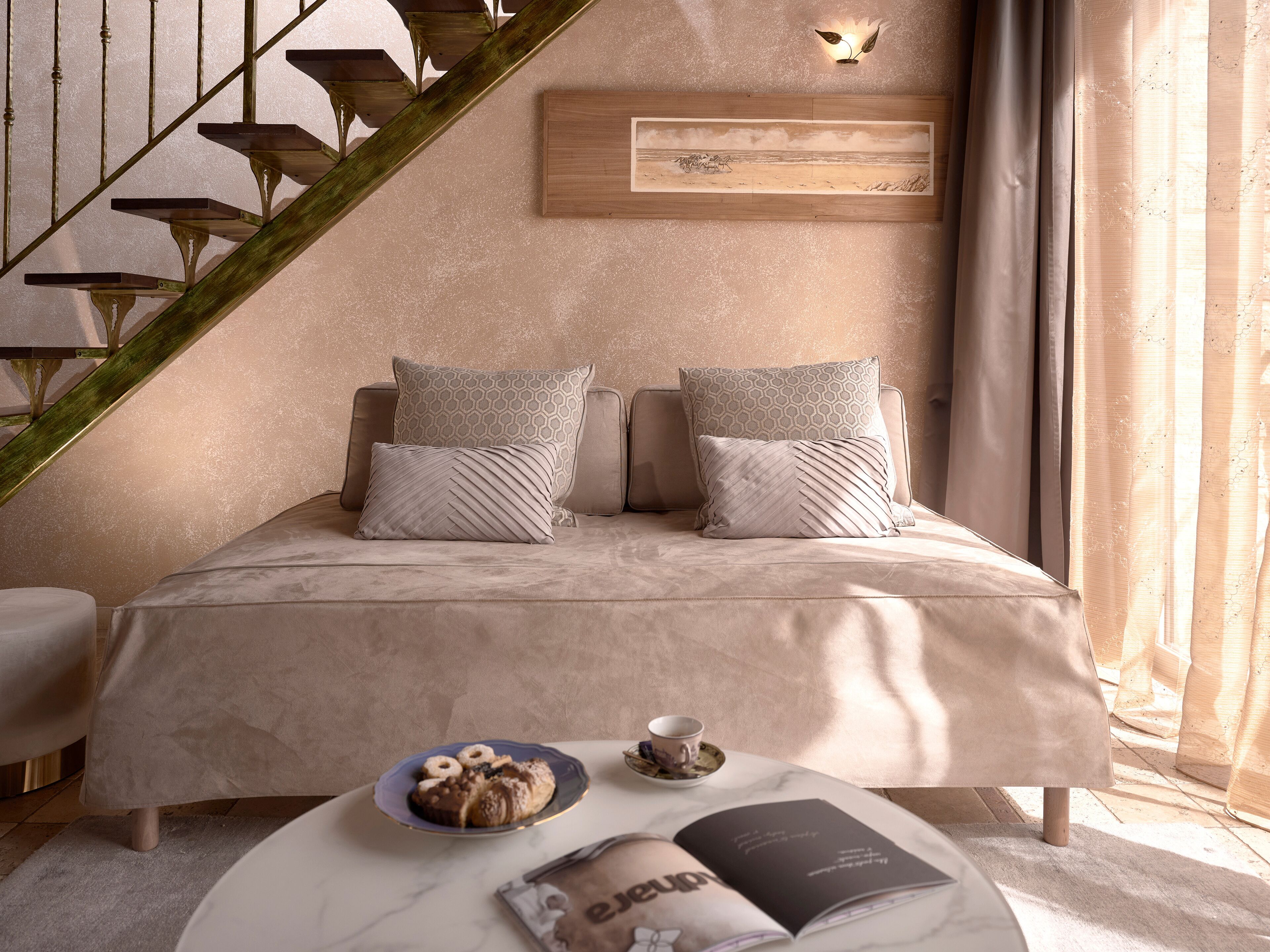 Un dormitorio con cama, almohadas, mesita de noche con una revista y un plato de pasteles, cabecera de madera, una obra de arte en la pared y una escalera.