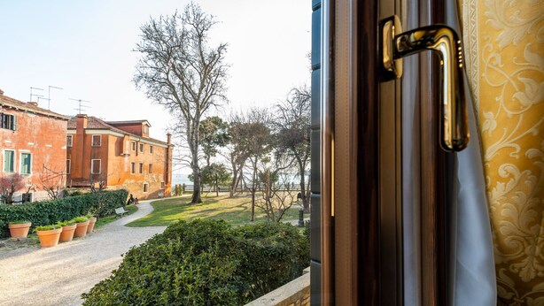 A view from a window with a brass handle, overlooking a paved path, a grassy area, and a building with red brick walls. - Apartment (Venezia)