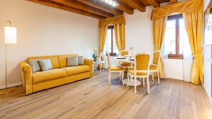 A room with a yellow sofa, wooden flooring, a dining table with chairs, and a window with curtains. - Apartment (Venezia)
