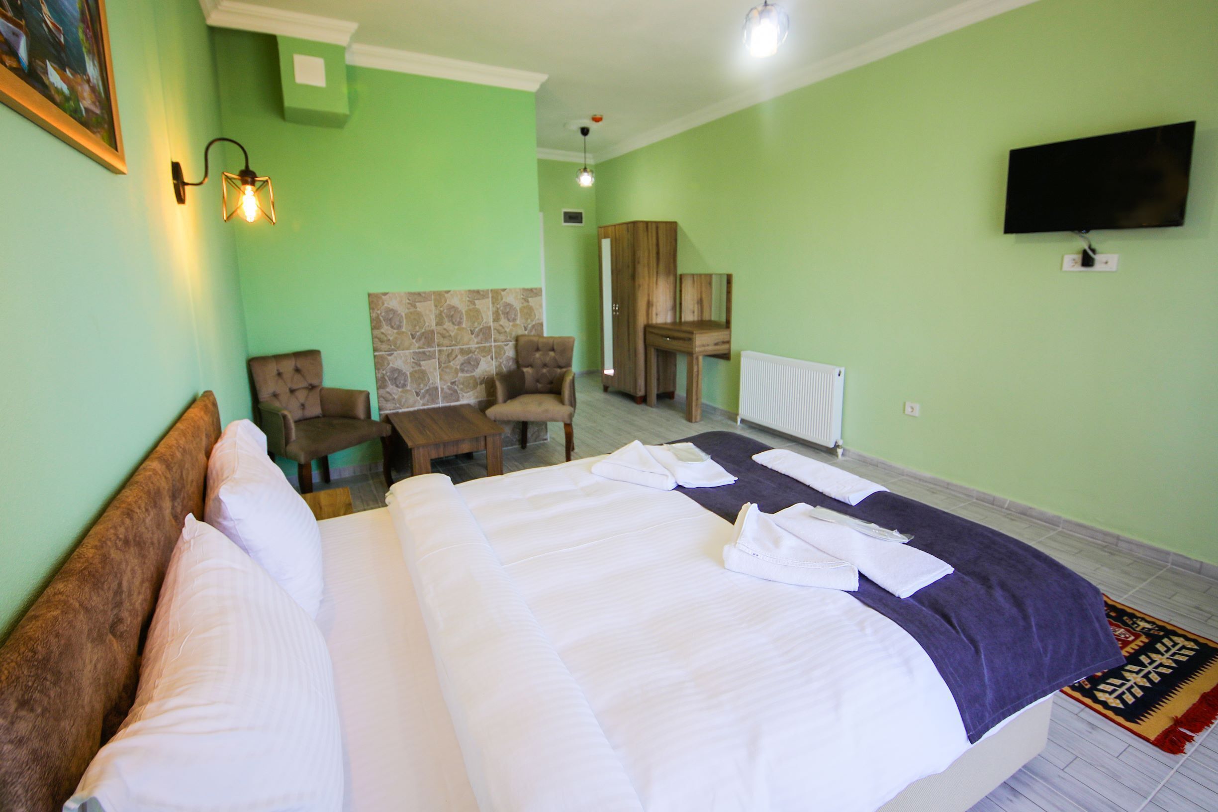Una habitación de hotel con una cama grande, cabecera de madera, zona de estar con sillas, una mesita, televisor montado en la pared y una alfombra estampada en el suelo.