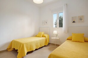 A room with two beds, a small table with a lamp, a window with curtains, and wall art.