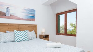 A bedroom with a bed, a nightstand, a lamp, a window, and a painting of a lighthouse. - House with Pool and Sea Views - Casa Can Divertit (Can Budella)