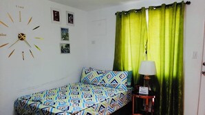 Une petite pièce, bien rangée, avec un lit, une horloge, une lampe et un rideau vert.
