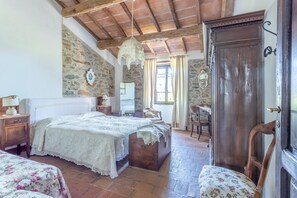 A rustic bedroom with a stone wall, wooden ceiling, a bed with a lace bedspread, a wooden chest, and a chair. - Apartment Il Tiglio with Shared Pool, Garden & Wi-Fi (Magliano in Toscana)