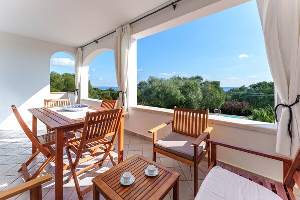 A terrace with wooden furniture, a table with cups, and a view of greenery and a pool.
