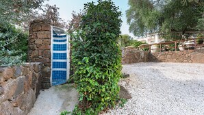 A stone wall with a blue-tiled gate, a gravel path, and a lush green hedge. - Holiday Apartment 'mamuthones' with Sea View, Wi-Fi, Terrace & Shared Pool (Orosei)