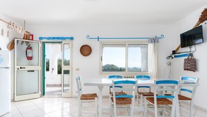 A dining area with a white table and chairs, a fire extinguisher, a television, and a window with a view. - Holiday Apartment 'mamuthones' with Sea View, Wi-Fi, Terrace & Shared Pool (Orosei)